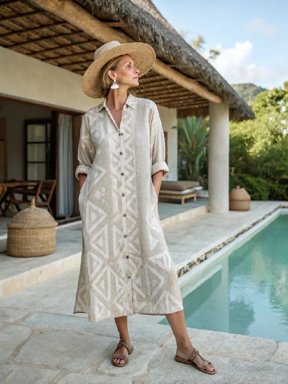 Woman in a patterned dress and straw hat standing by a poolside with a thatched-roof structure in the background.