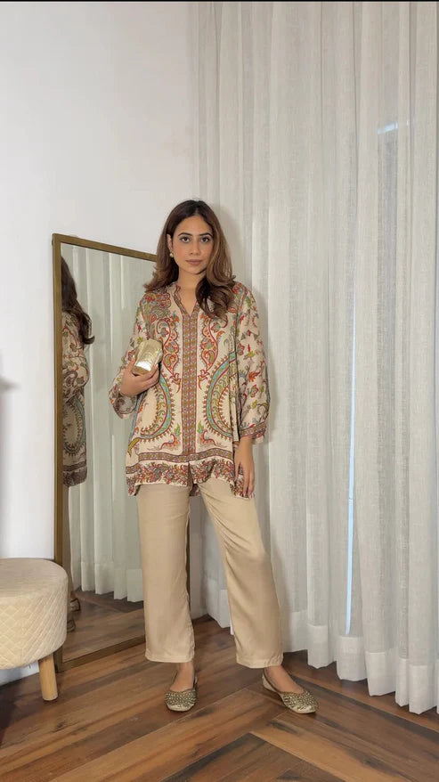 Woman standing in front of a mirror wearing a patterned top and beige pants, holding a phone.