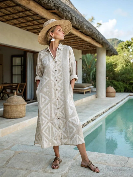 Woman in a patterned dress and straw hat standing by a poolside with a thatched-roof structure in the background.