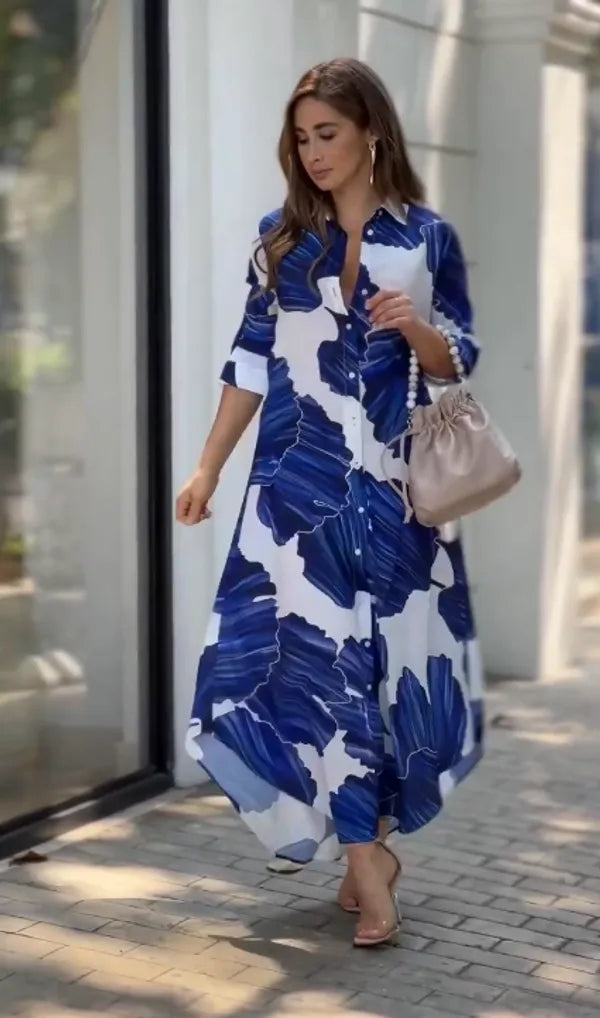 A woman wearing a long maxi dress with blue floral prints, paired with a beige bag and sandals, standing on a sidewalk.